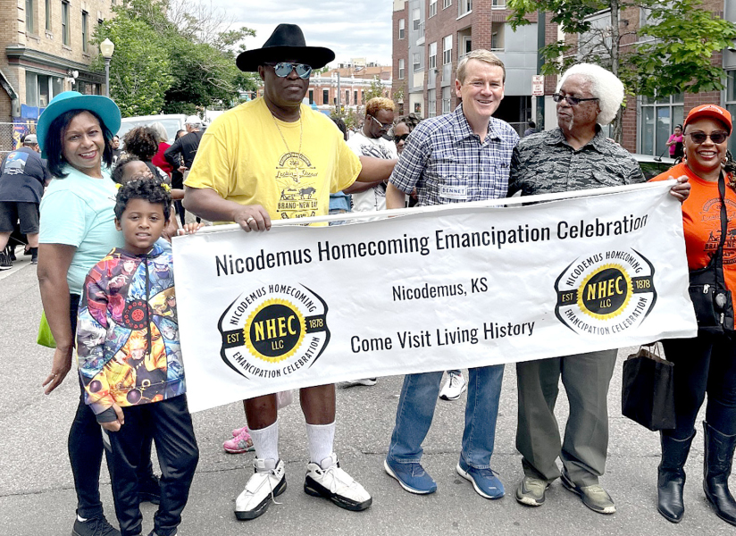 NICODEMUS DESCENDANTS from Colorado in the 2023 Denver Juneteenth Parade representing the Nicodemus Homecoming Emancipation Celebration with their banner included from left: Loval VanDuvall, Karmen Robinson holding grandchild Amira Wilson, Orval VanDuvall, United States Senator Michael Bennet from Colorado, Virgil (Poncho) Robinson and Linda Jones.