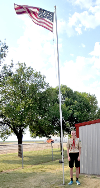 RILEY ROGERS stands proudly next to a completed flagpole, with the flag waving at the Plainville ballfields.