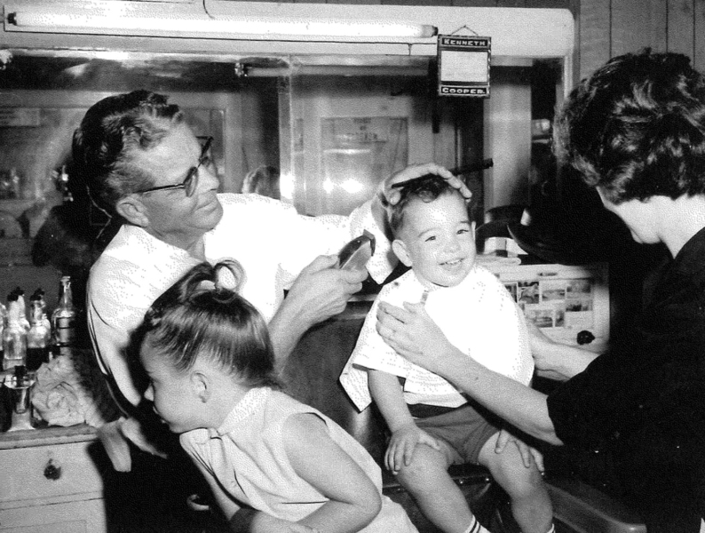 THIS STORY REMINDED THE HAMILTON FAMILY of a couple of photos they had of the Cooper Barber Shop. In this photo, dated August 12, 1965, Kenneth Cooper is giving Bobby Hamilton his first haircut. The photo shows Marlene Hamilton (mother) trying to keep Bobby from squirming while his sister, Susan, age 4, is distracted by Cooper’s barber tools.