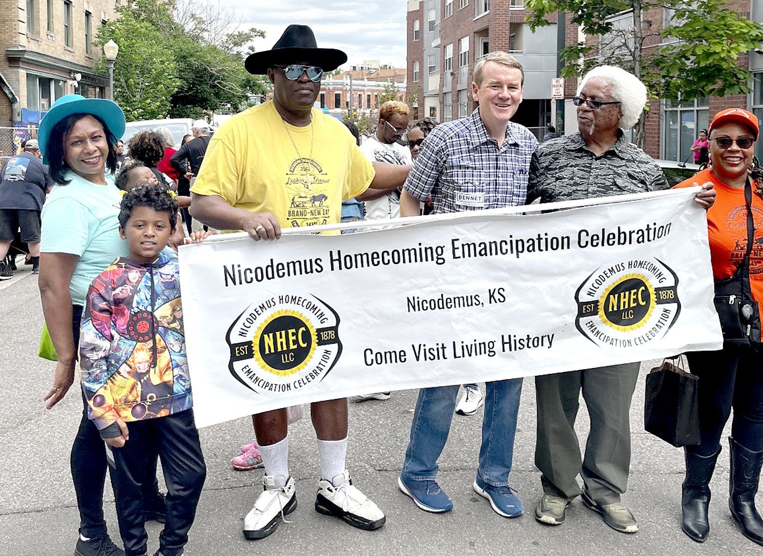 NICODEMUS DESCENDANTS from Colorado in the 2023 Denver Juneteenth Parade representing the Nicodemus Homecoming Emancipation Celebration with their banner included from left: Loval VanDuvall, Karmen Robinson holding grandchild Amira Wilson, Orval VanDuvall, United States Senator Michael Bennet from Colorado, Virgil (Poncho) Robinson and Linda Jones.